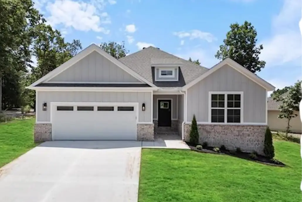 Newly built earth-tone ranch home with board-and-batten siding and a stone skirt on a wooded Bella Vista lot