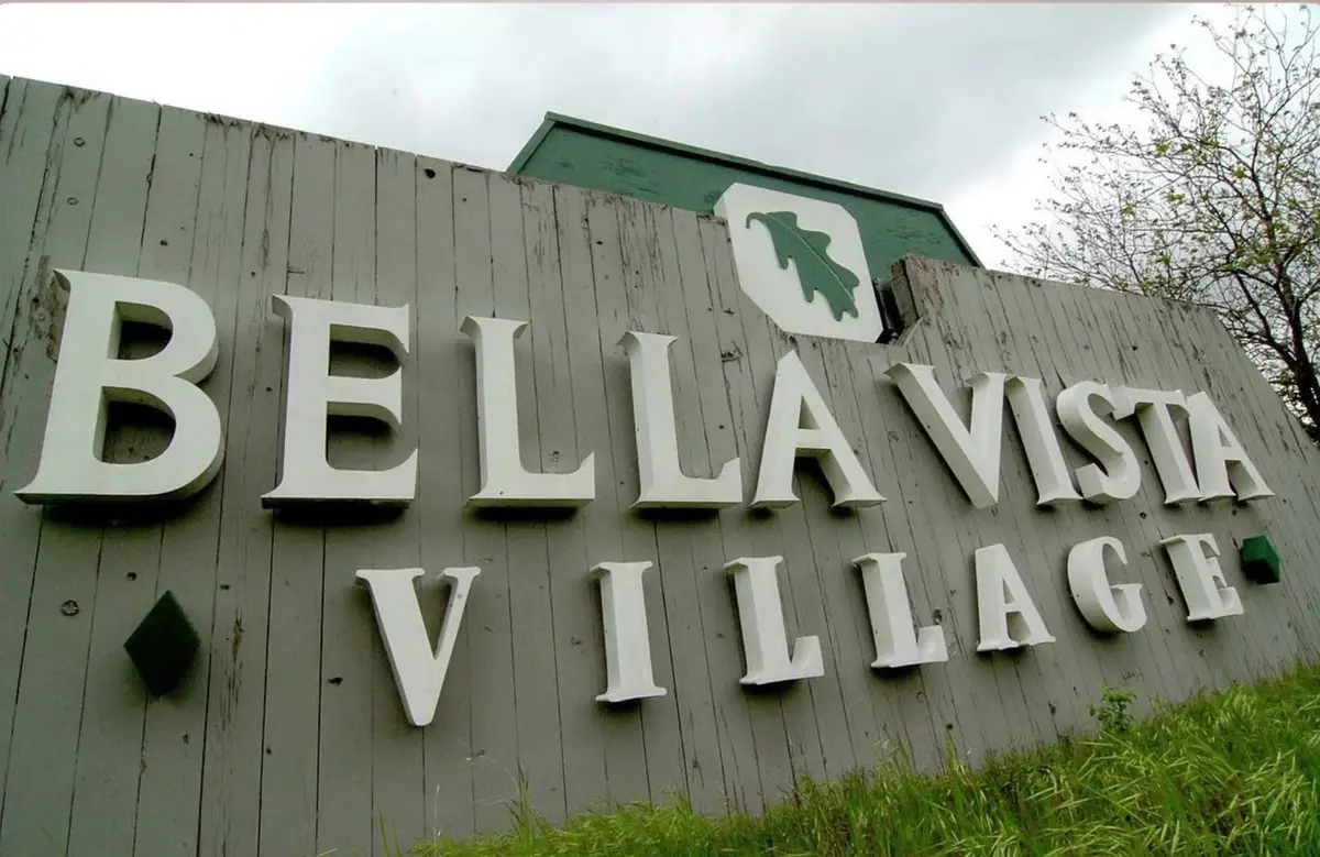 Weathered wooden Bella Vista Village entrance sign with an oak-leaf emblem in Northwest Arkansas