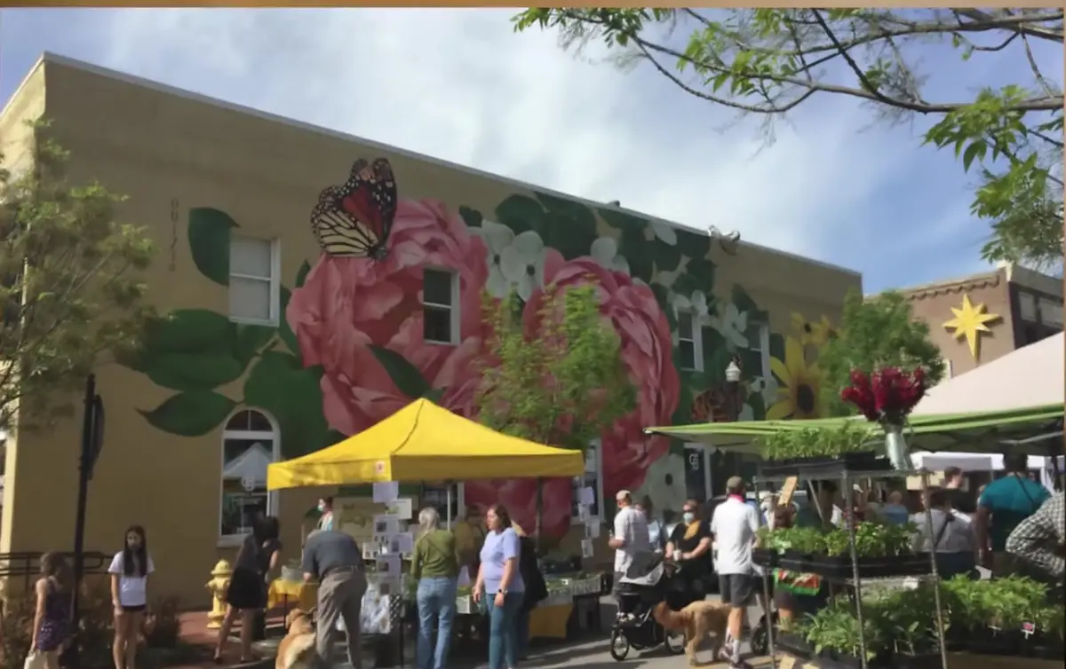 Shoppers and vendor tents at the Bentonville Farmers Market on the downtown square in Arkansas, beside a large floral mural