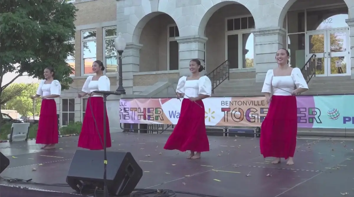 Four dancers in white blouses and red skirts performing on an outdoor stage at a Bentonville community festival