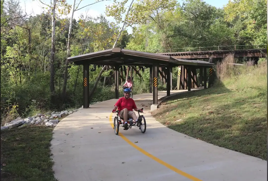 Cyclist riding a recumbent trike on a paved shared-use greenway trail beneath a covered shelter in Fayetteville, Arkansas