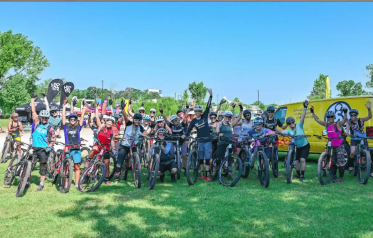 A large group of mountain bikers raising their hands together at a trailhead in Northwest Arkansas