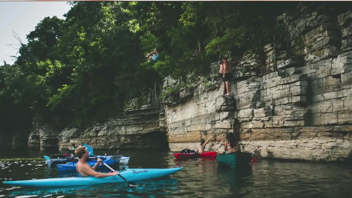Kayakers paddling below a limestone bluff on an Ozark lake in Northwest Arkansas as a swimmer jumps from the rock ledge
