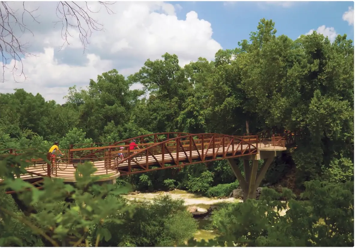 Cyclists crossing a weathering-steel pedestrian bridge over a wooded creek on a Northwest Arkansas trail