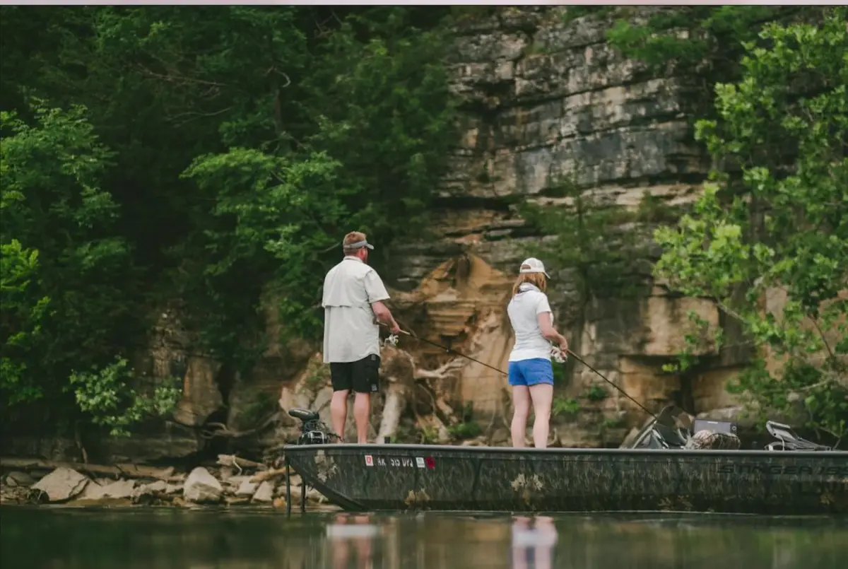 Two anglers fishing from a boat beneath a wooded limestone bluff on an Ozark lake in Northwest Arkansas