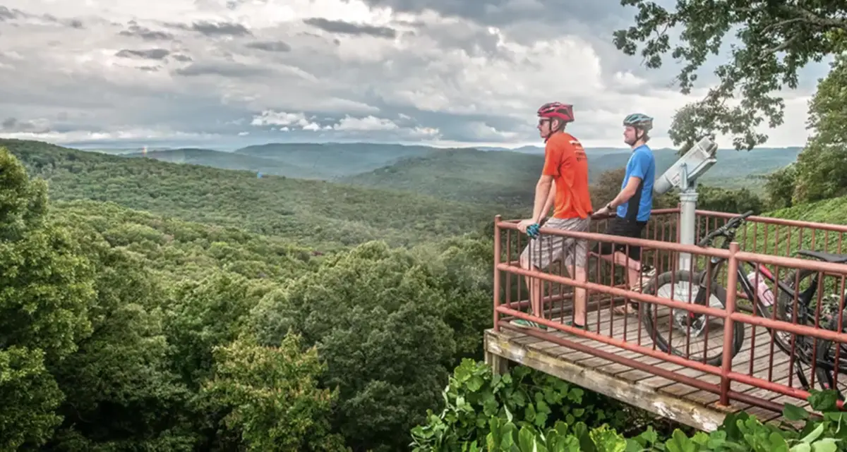 Two mountain bikers resting at a wooden overlook deck above forested Ozark hills in Northwest Arkansas